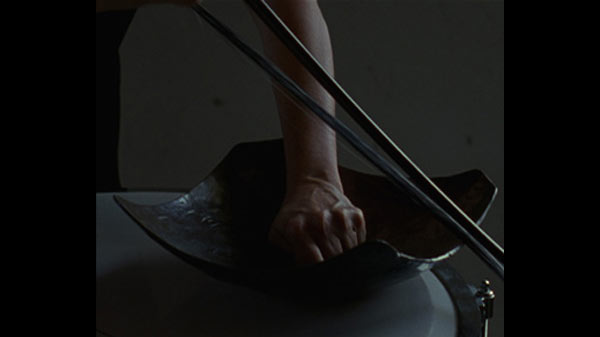 An arm is pushing its fist against a bottom of a square-shaped bowl. The accentuated sinews on the fist indicate the amount of force that is generating. A bow of bass is held above the bowl. Only rims of the bowl and edges of the bow are reflecting light in a very dim space.