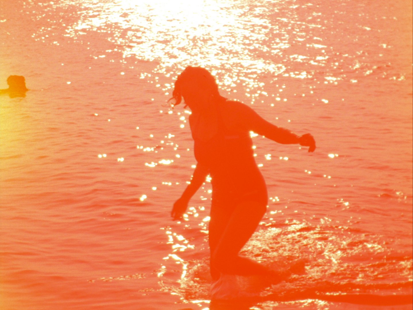 A washed out reddish image of a woman in the water with the sun shining, a film still from Pyramid by Margaret Salmon