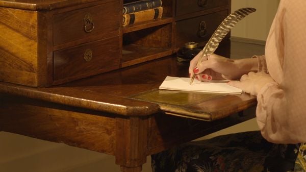A person in a pink blouse sitting at a wooden desk. The focus is on a hand with red fingernails writing with a feather quill