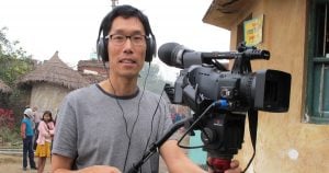 An East Asian man operating a camera on a tripod at an outdoor set. He is wearing a large headphones and wearing a grey t-shirt.