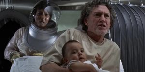 A agitated man holding a baby on a hospital bed which is pushed by a woman in a futuristic protective suit.