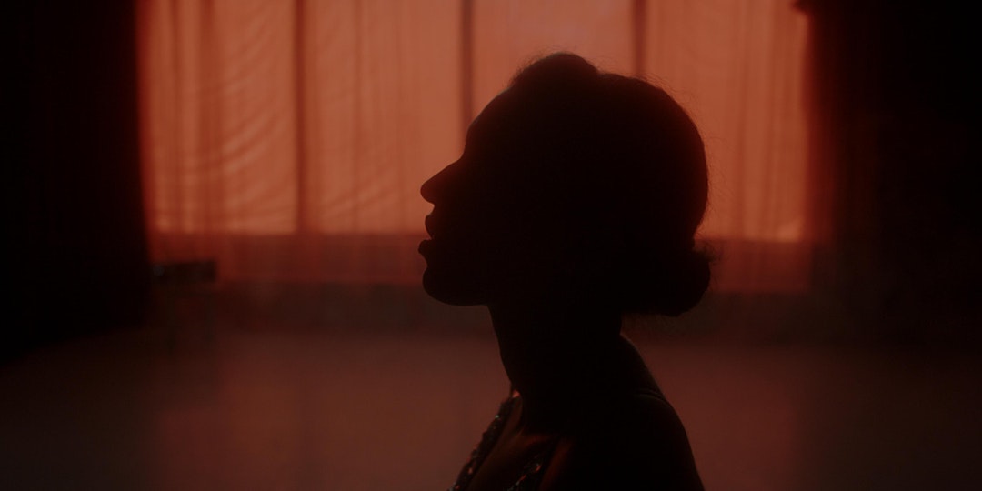 A silhouette of a person stands in front of warm atmospheric lighting coming through the translucent window curtain.