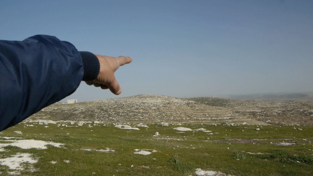 Colour photograph of a landscape showing a clear blue sky, grass and clusters of rocks in the distance. There is an arm visible in the frame from the left side of the image. Dr. Ibrahim Mekharzeh, who is dressed in a blue sleeved jacket, is gesticulating and pointing at significant archaeological sites in the distance (southern West Bank).