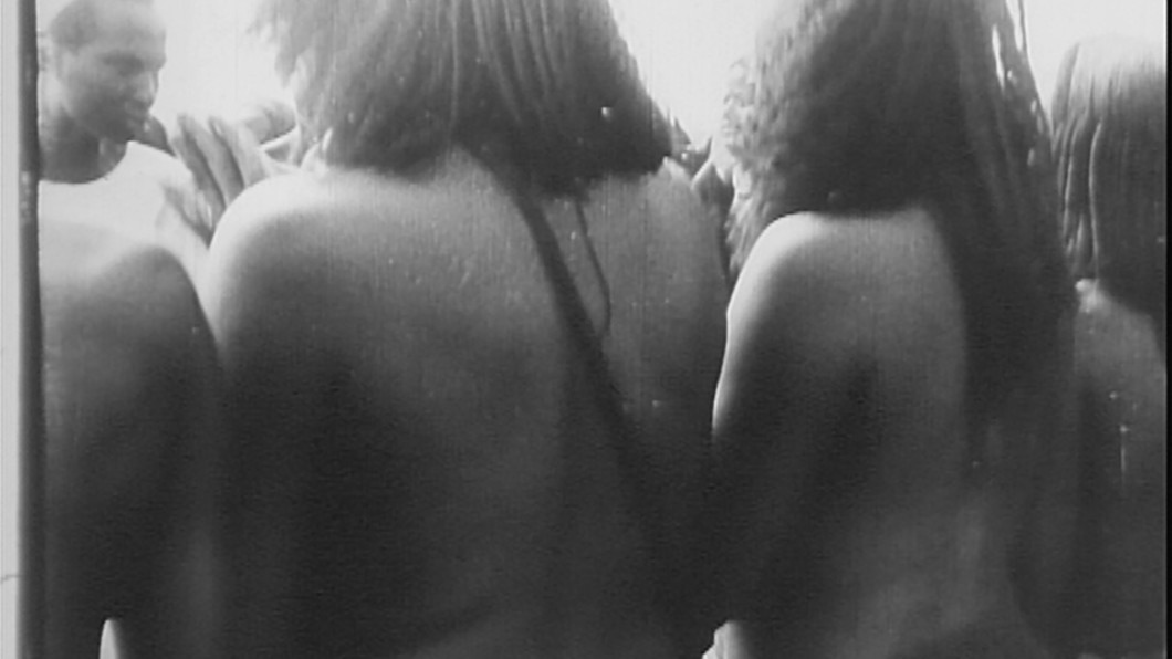 The backs of naked black women standing in a row. A face of a black man in front of them is seen at the left top corner.
