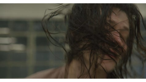 A still from a film Bathing by Patrick Staff. A blurred image in motion of a person’s face swathed with one’s long hair . The background is gloomy and out of focus. 
