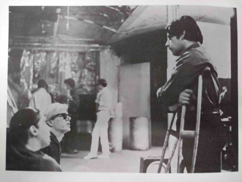 In a printed B&W photograph a man is standing and leaning on crutches. Two people sitting are looking up and speaking to the man standing.