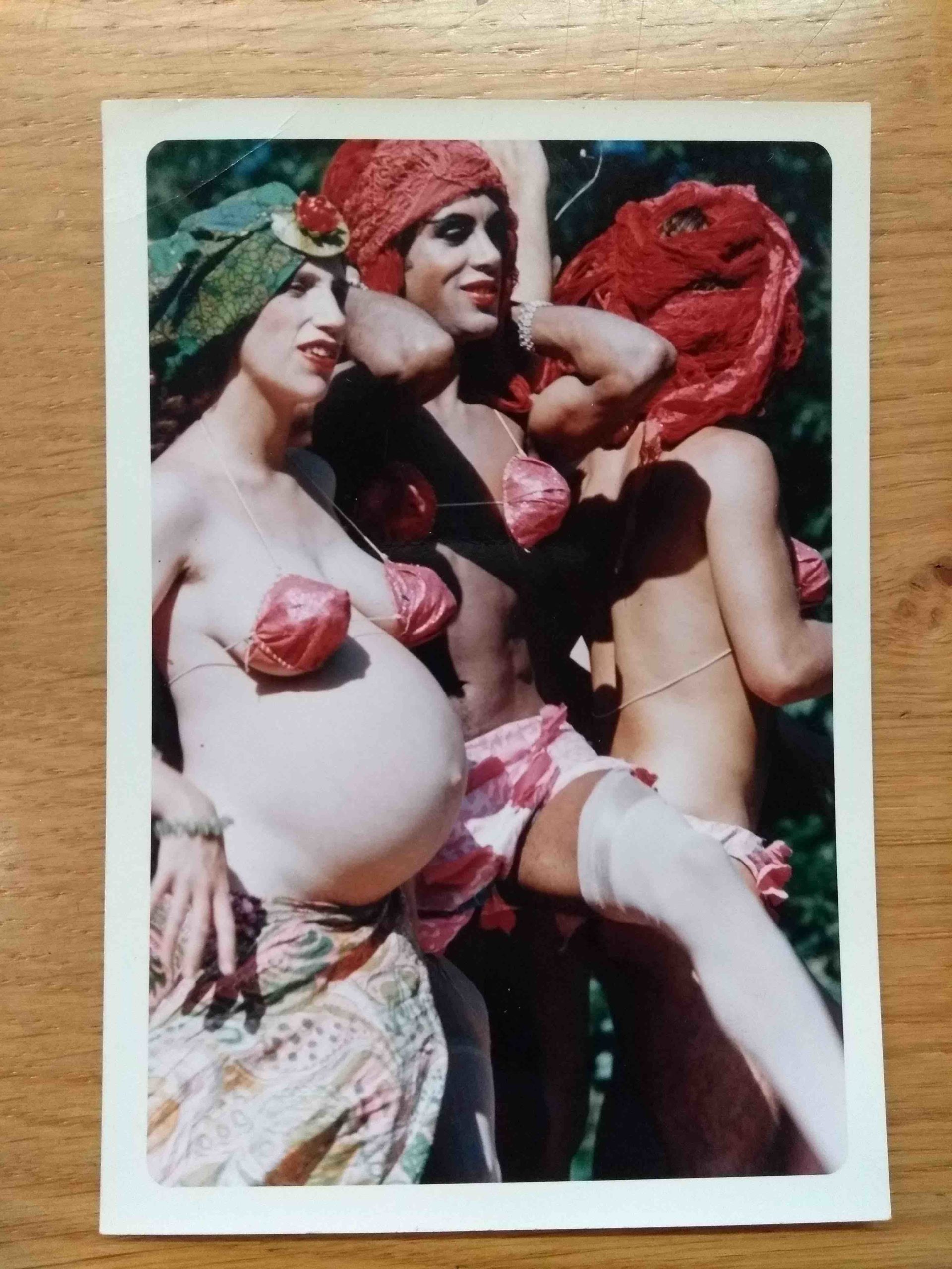 A color photograph placed on a wooden surface. The photo captures three bodies all in bikini tops and elaborate head pieces. The one on the very left is heavily pregnant