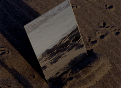 A piece of mirror is mounted on a sand beach at 45 degree angle. The mirror reflects the sea shimmered in the sunlight and small mounds of sand. 