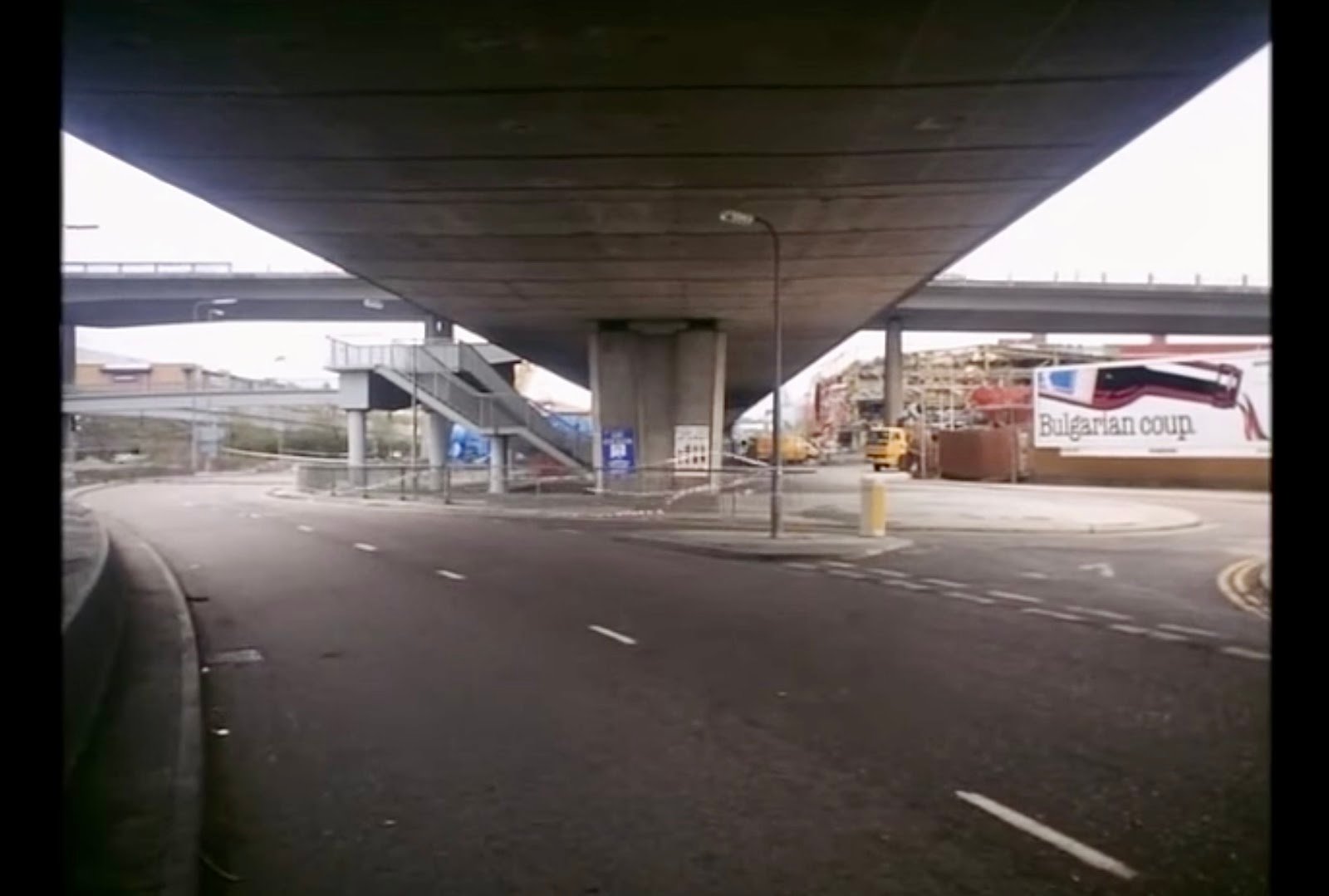 A view of an empty underpass under which A road diagonally cuts through. Outlines of a bridge create a vanishing point at the centre of the frame.