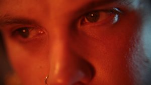 A closeup of a white person’s eyes looking intensely at something outside the frame. A dark orange light hues the face and there is a hint of a blue light coming from behind.