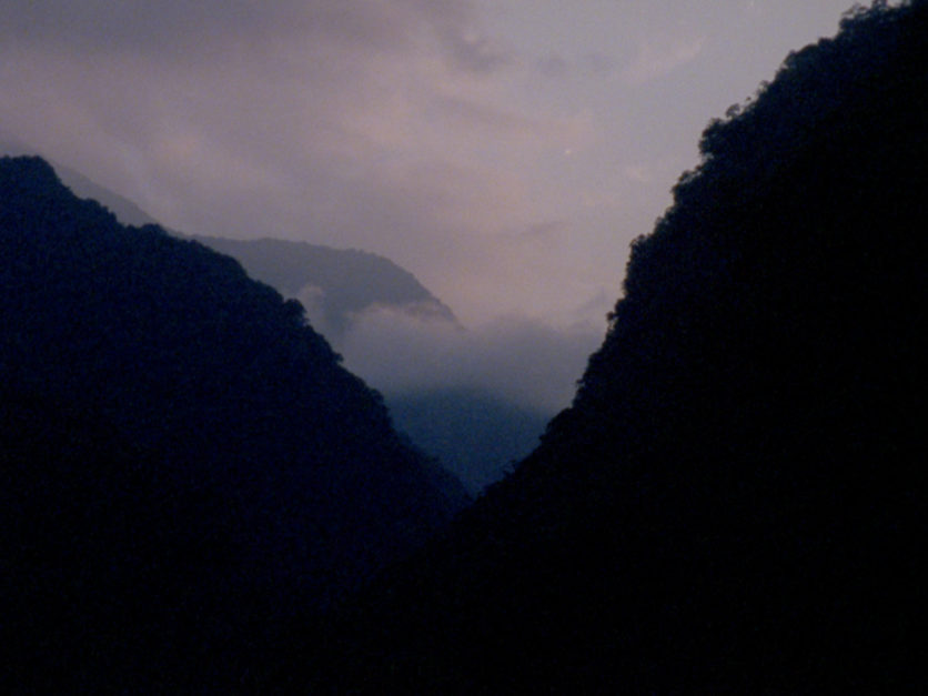 Silhouettes of Massive mountain slopes create a v shape through which purple-hued clouds and sky are viewed.