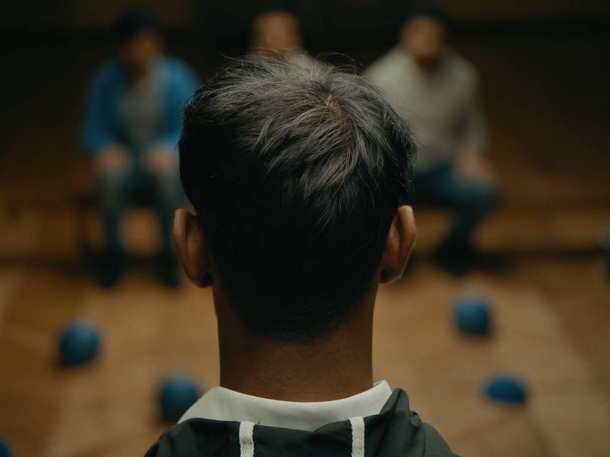 A close-up image features the back of the head of young dark skinned man in the foreground. In the background, three other men, out of focus, are facing him. They are seated in what appears to be a gymnasium with a wooden floor. Bright blue balls are scattered across the floor.