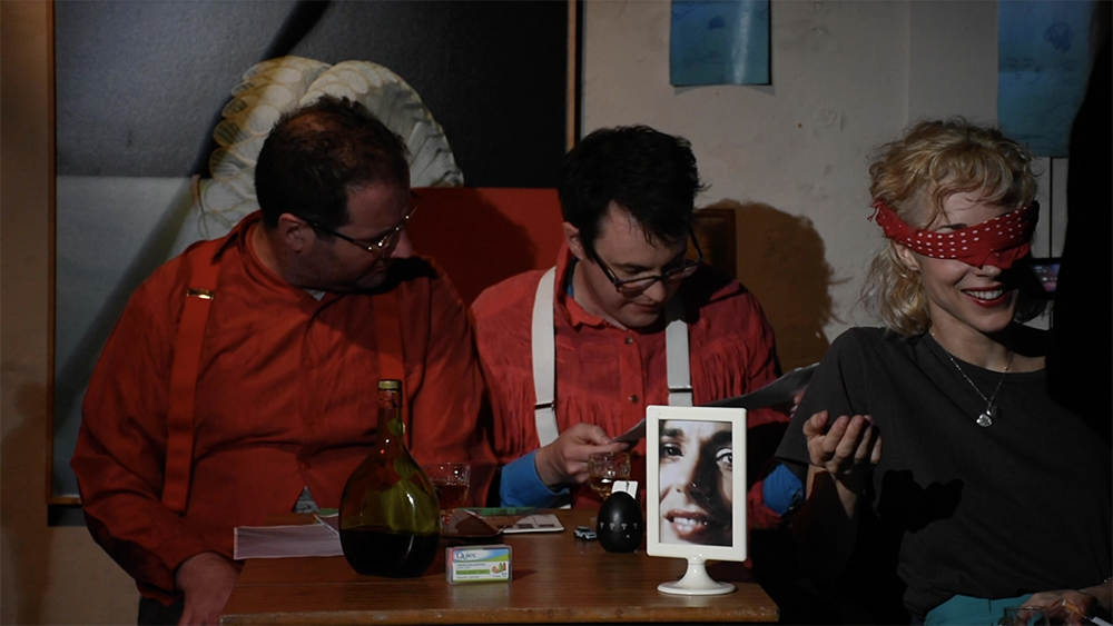 Two white men in red shirts are looking at a piece of paper together. In front of them is a table with a wine bottle and glasses as well as a picture frame filled with a zoomed in photo of a white woman. On the right is a white blond woman whose eyes are covered with red clothes, smiling. 