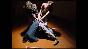 Two women are pulling a man’s waist up while their upper body creates V-shape. The man’s head and feet are on the floor creating a reversed V-shape. a pair of crutches fell on the orange floor.