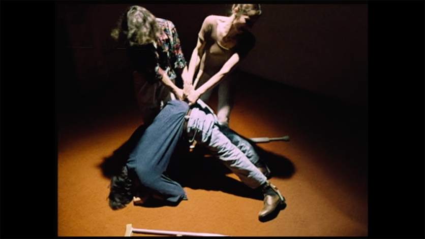 Two women are pulling a man’s waist up while their upper body creates V-shape. The man’s head and feet are on the floor creating a reversed V-shape. a pair of crutches fell on the orange floor.