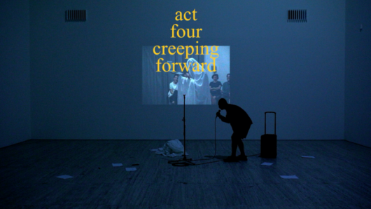 A white cube space is dimly lit by blue-hued light. A silhouette of a person speaks to a mic with a hunched back. An image of a person covered with a shite sheet in a kind of ceremony is projected on a wall on which is overlayed with a large caption that writes “act four creeping forward”