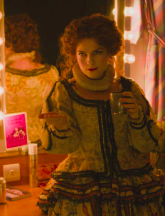 A red haired woman in a Victorian dress costume stands in front of a mirror in a backstage powder room. She holds a copper cup and stares at someone with a smirk on her face. 