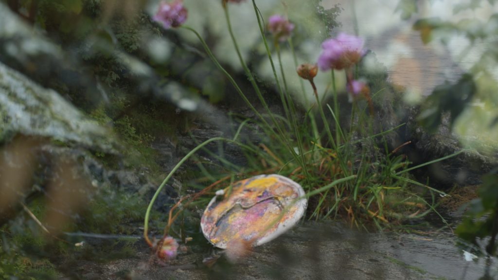 A colourfully painted ivory pendant is abandoned on a wet ground. it seems to lean on a bunch of wild flowers
