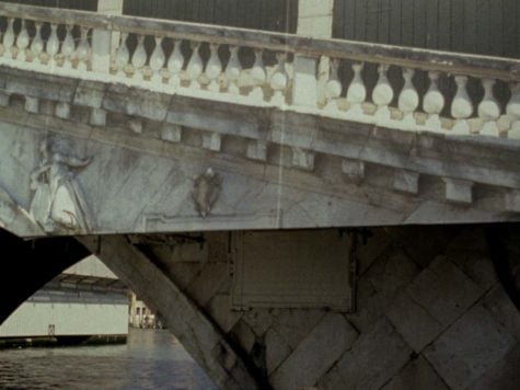 A view of a bridge cropped from a top of a pile built with wedged stones to a deck slab decorated with a figure and railing. 