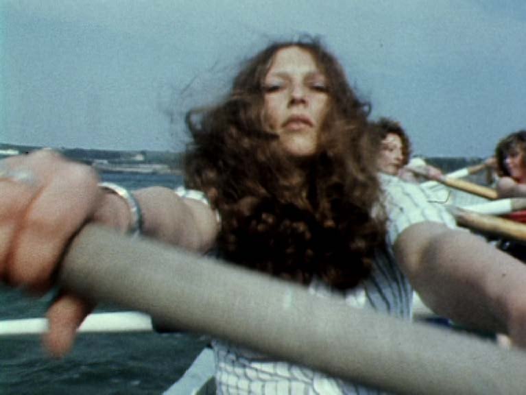  A white woman with voluminous curly hair rows in a boat with her arm fully stretched forward. Behind her are two women rowing side by side. 