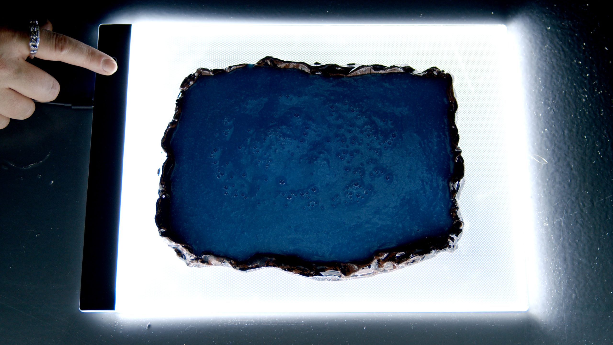On a glowing led lightbox is a ceramic tray with erratic edges filled with blue liquid. A finger wearing a silver ring touches the power button on the lightbox