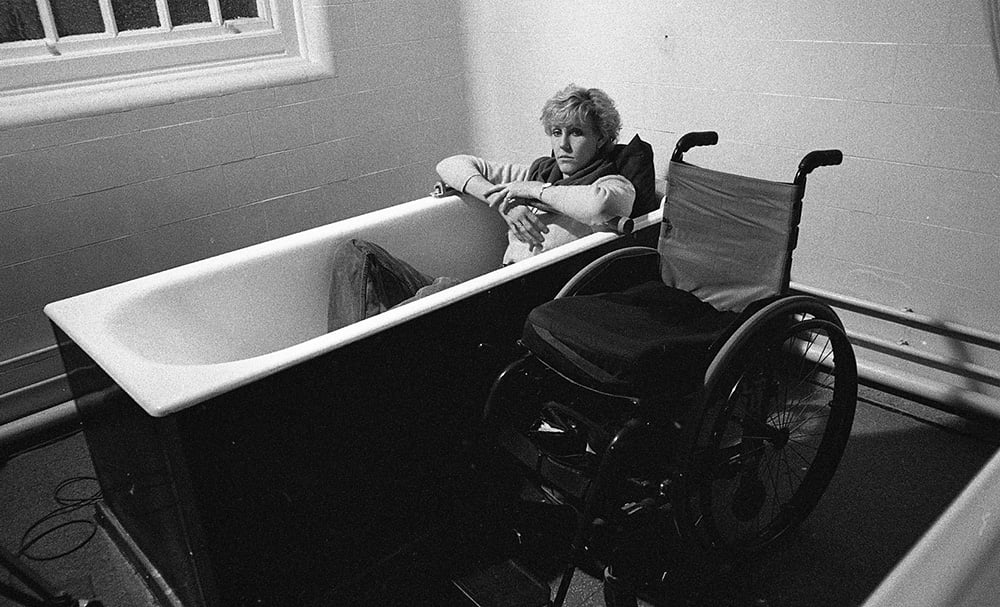 Ellen Stohl, a white woman with a short cut is sitting in a bathtub. A wheelchair stands next to her.