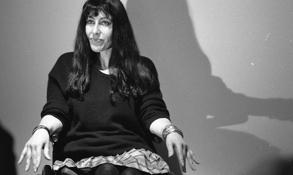 A portrait of Julie Umerle. A B&W photograph of a white woman with a long black hair sitting on a chair. A harsh spotlight on her creates a distinct silhouette on the wall behind her. 