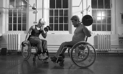 A white woman and white man in wheelchairs weightlifting in a well-lit indoor space.