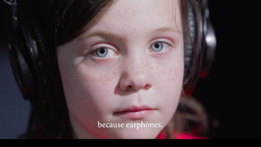 still image from Receiver by Jenny Brady a young white girls face in close up wearing headphones, she gazes calmly into the camera, there is a caption in white which reads 'because headphones'