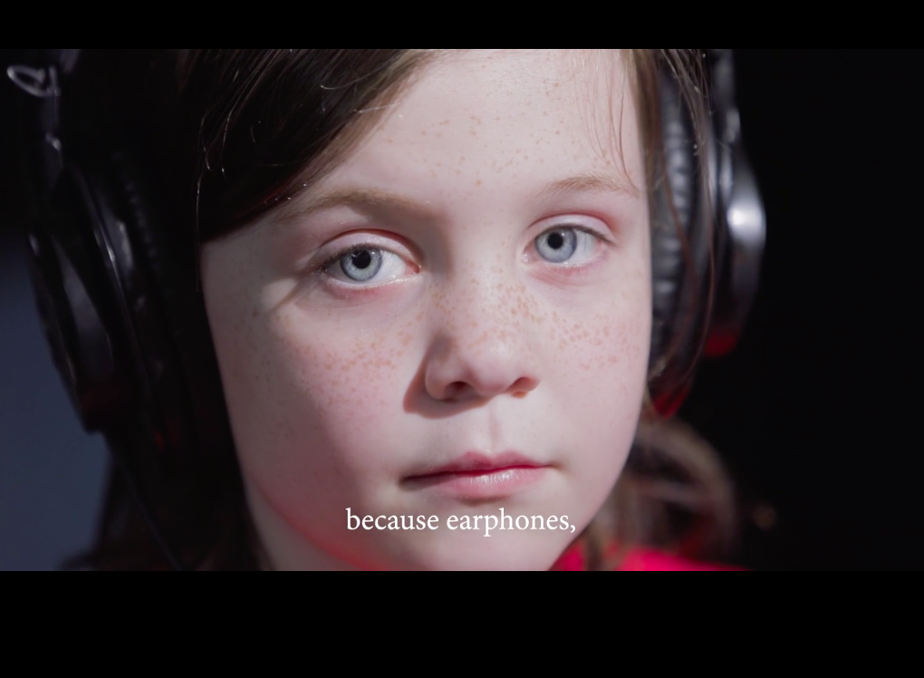 still image from Receiver by Jenny Brady a young white girls face in close up wearing headphones, she gazes calmly into the camera, there is a caption in white which reads 'because headphones'