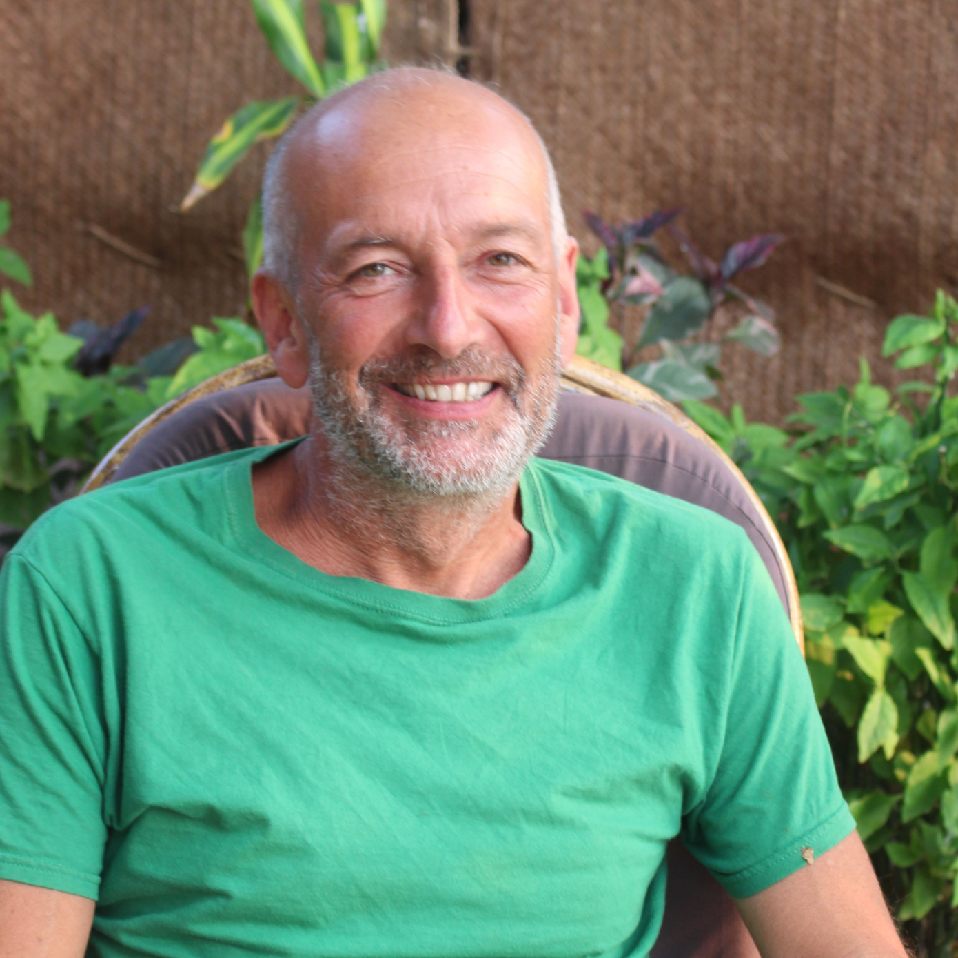 portrait of George Barber, a middle aged white man with a bald head and grey beard wearing a green t-shirt, smiling and sitting in front of some plants