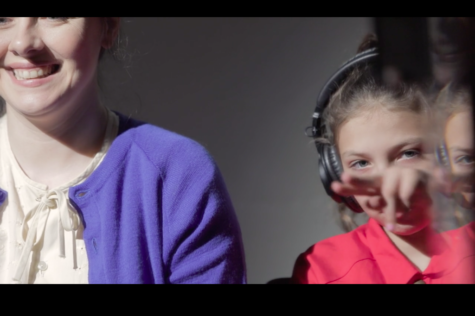 still image from Receiver by Jenny Brady, an older white woman and white girl who is wearing headphones sit next to each other. The woman smiles off camera while the girl signs, her hand in front of the image, there is some mirroring off the girl on the right hand side of the image