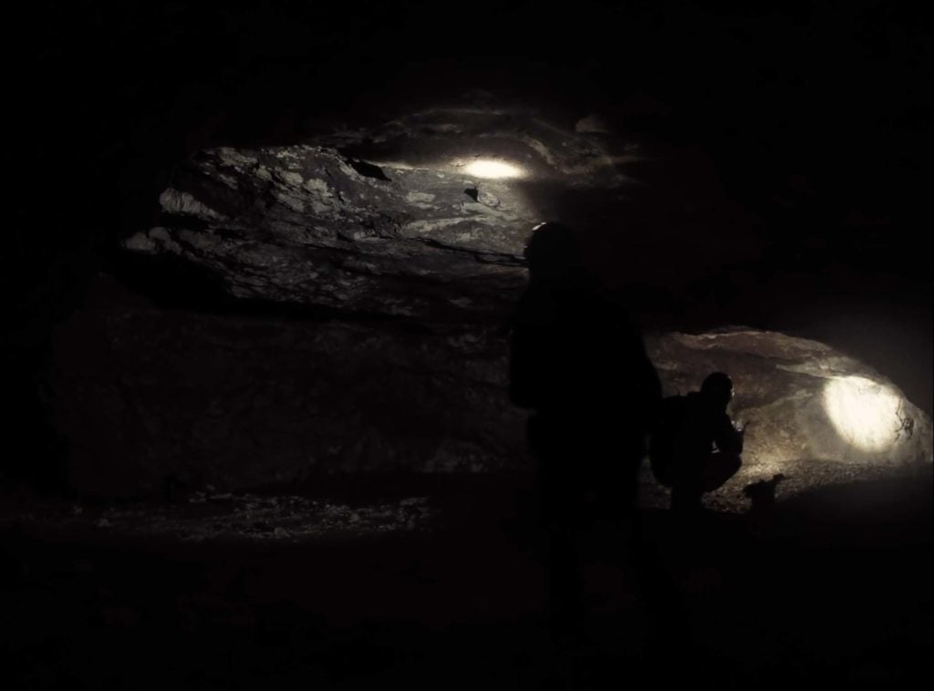 Inside a dark cave. Two people illuminate parts of the surface revealing rocky texture with flashlights. 