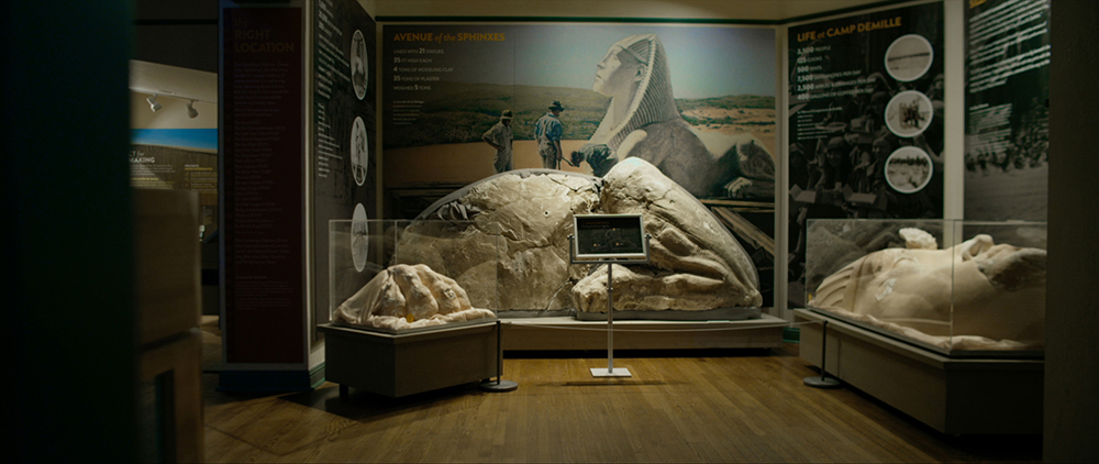 A museum display of three dismantled pieces of sculpture. On the wall is a picture of a Sphinx and three archaeologists in front of it. 