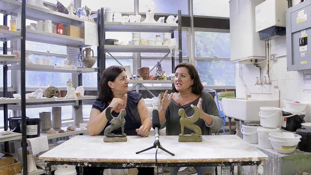 In a lab filled with models, paint buckets, ancient objects, two women talk in front of a mic on a workstation covered with white paints. A rusty sculpture of half human and half bird sits on the left and a seemingly replica sits on the right on the table. 