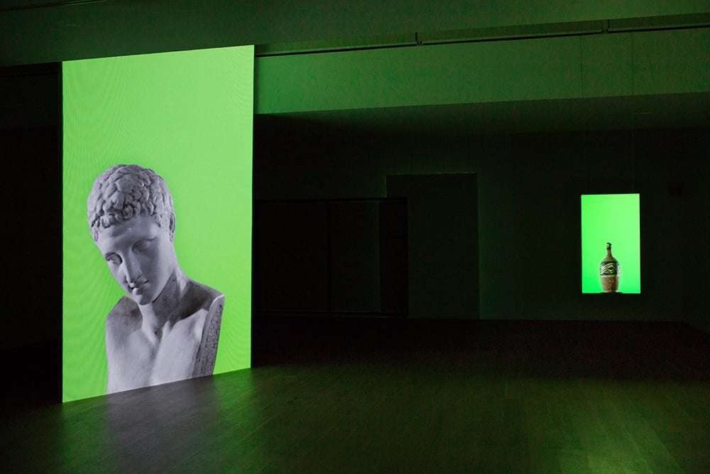 An installation view of two channel video projections in a dark gallery space. On both projections objects are set against a bright green background. The left is a marble bust and the right is an ancient vase.
