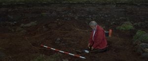 A white old man in a red jacket holds a small brush and sits in a pit on a demarcated archaeological site. He stares at a pile of rocks and soil near him.