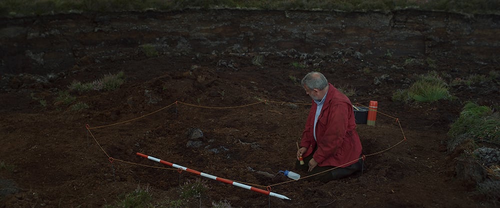 A white old man in a red jacket holds a small brush and sits in a pit on a demarcated archaeological site. He stares at a pile of rocks and soil near him.