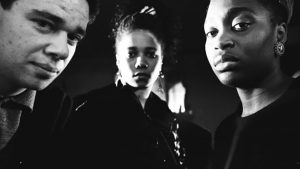 Three people, a white man and two black women, looking directly into the camera under a dramatic lighting creating a film noir look.