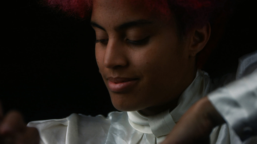 A black person cropped from the forehead to shoulders, wearing a silk blouse, against a black background. THe person is looking down with a subtle smile. A glimpse of a red afro on the top edge of the frame. 