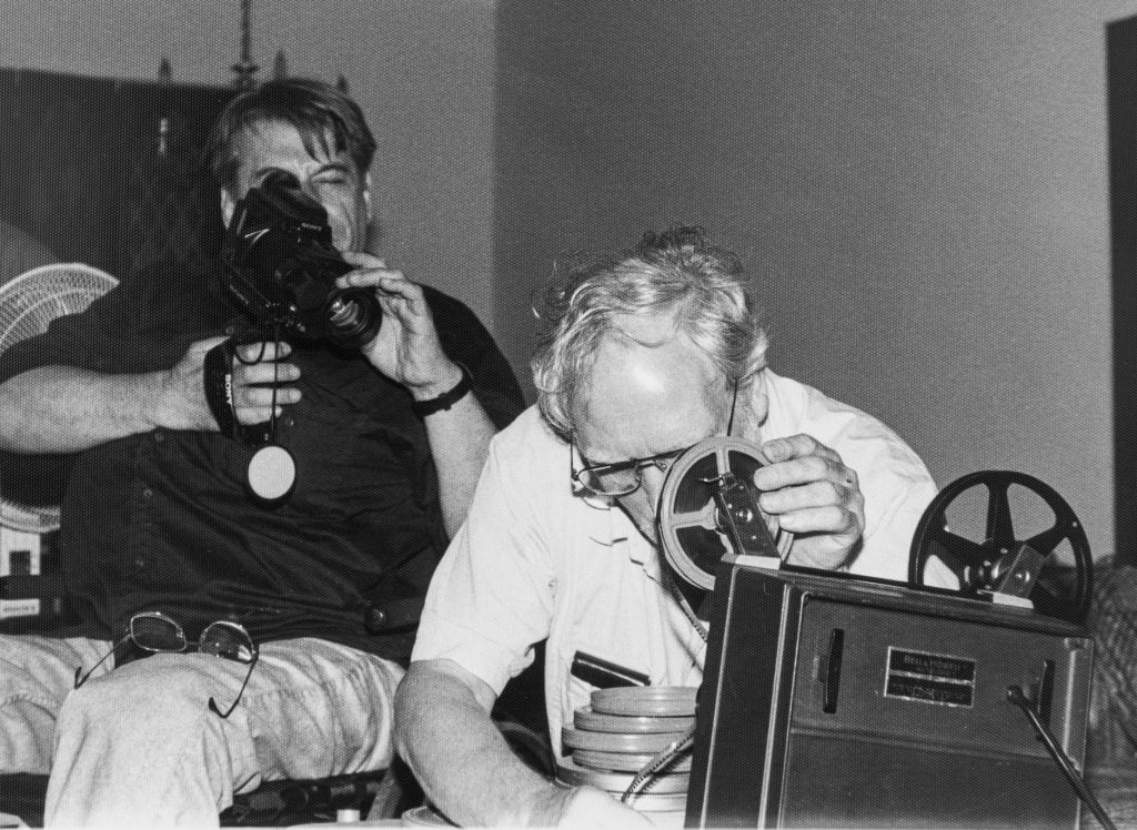 Two white men. The younger one on the left holds a camera and looks through a viewfinder. The older one on the right is crouched behind a film projector, holding a small reel of film near his face. 