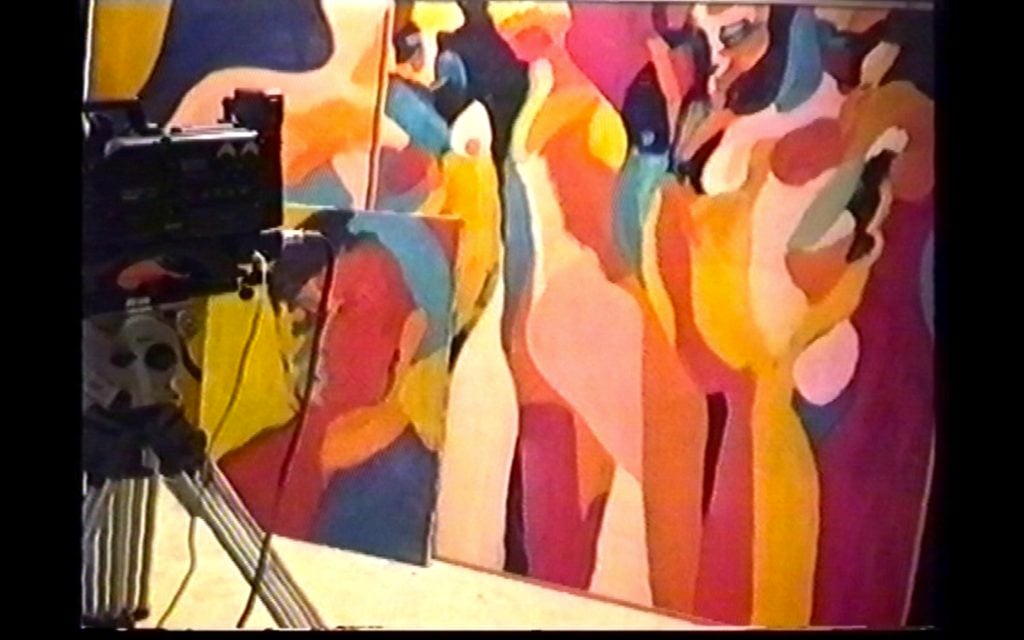 A video camera mounted on a tripod is filming colourful abstract paintings of human figures leaned against the wall. 