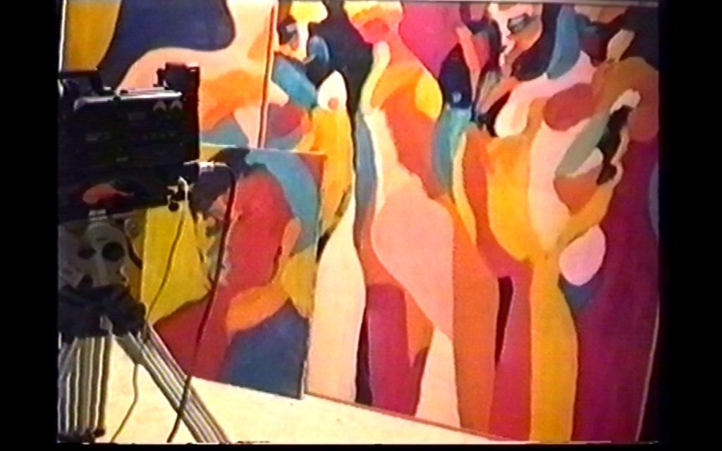 A video camera mounted on a tripod is filming colourful abstract paintings of human figures leaned against the wall. 