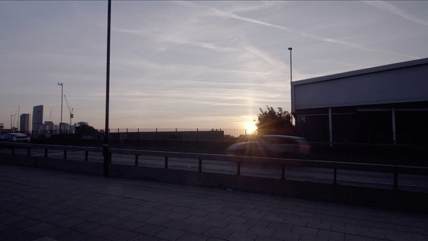 A road that leads to a bridge against a backdrop of a grey sky and the beaming sun at dusk. A SUV passes in a blur. Highrise buildings and cranes appear to be very small in the background.