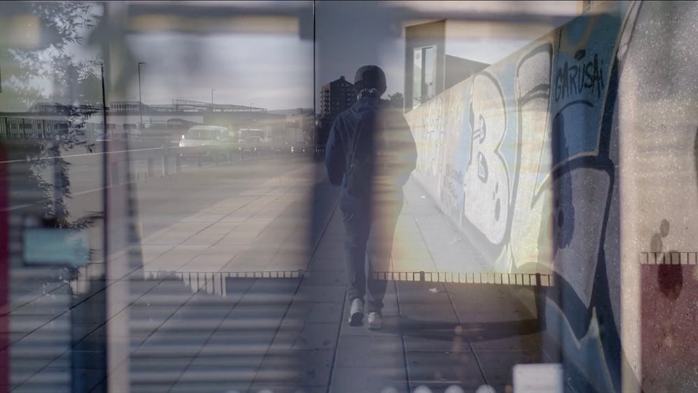 A person with a backpack walks on the pavement between a road with cars and a wall of graffiti. The image is overlaid with a river reflecting sunset through windows. 