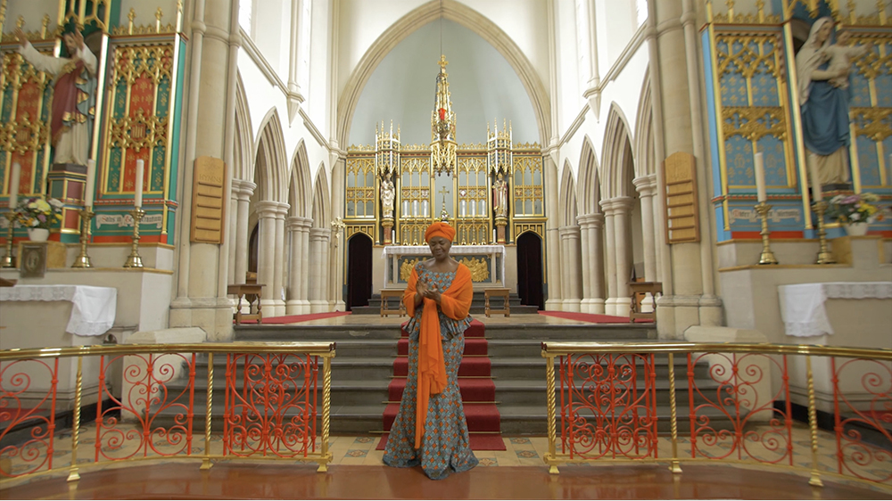 A black woman wears a turquoise and red patterned dress and bright orange headwrap and scarf. She stands in front of an altar decorated with statues and golden structures. 