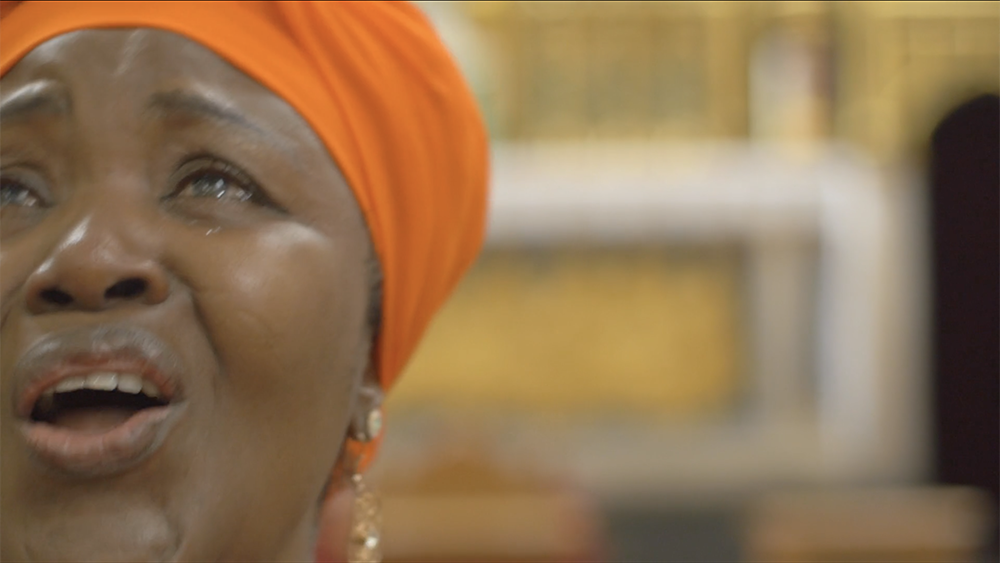 A close look at a black woman with an orange headwrap, who seems to be singing. Tears rolling on her chick and the background is very blurry and indiscernible. 