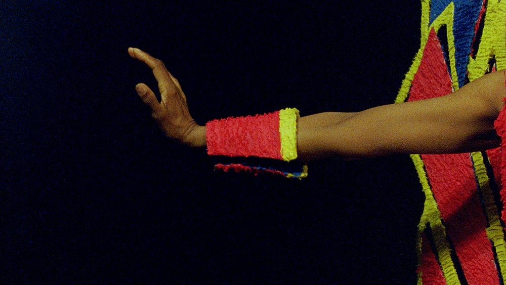 A dark-skinned person’s arm stretches out from the right side of the frame against a pitch black background. The arm wears a thick neo red and yellow bracelet and we see a glimpse of a neon red and yellow costume on the right. 