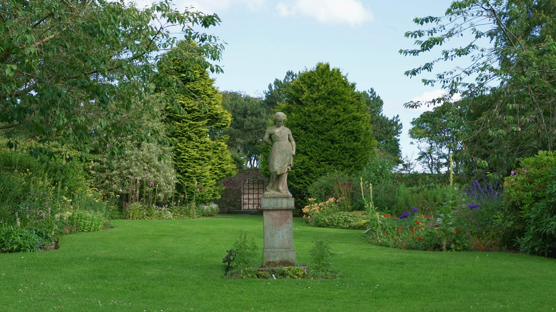 A beautifully cultivated garden. At the centre is a statue of a woman on a podium.