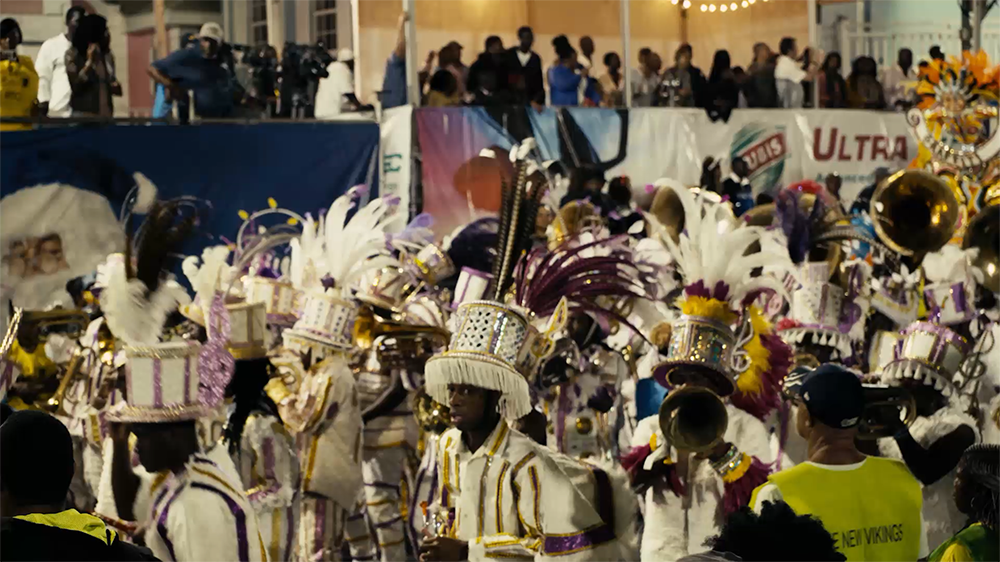 Marching band consists of black performers in white uniforms decorated with feathers. Behind them is a group of people observing the parade. 